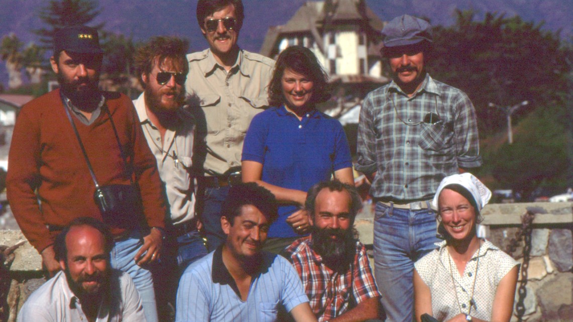 A group of men and women in 1970 clothing pose together outdoors.